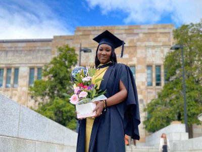 A young African woman stands in graduation cap and gown and holding flowers outside 喵喵直播's sandstone Great Court.